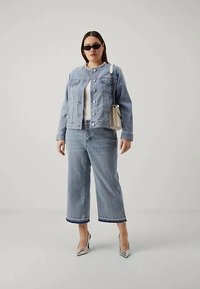 Nuori nainen, joka on pukeutunut vaaleansiniseen farkkutakkeeseen ja leveälahkeisiin farkkuihin, valkoiseen puseroon, hopeisiin korkokenkiin, mustiin aurinkolaseihin ja kantaa vaaleaa olkalaukkua.