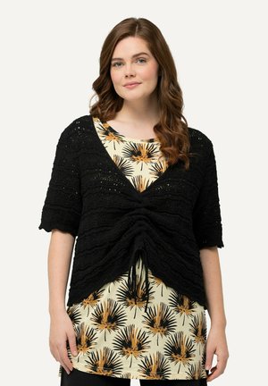 Musta neulecardigan, jossa on lyhyet hihat, teksturoitu muotoilu ja rypytetty etuosa. Yhdistetty kuviolliseen kermaiseen toppiin, jossa on palmulehtiä.