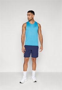 Kék sportos ujjatlan felső fekete logóval, navy színű sorttal párosítva. A modellen fehér zoknik és sportcipők találhatók, a szabott, illeszkedő dizájn kiemelkedő.