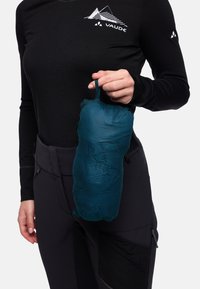 Sac de couleur sarcelle, pliable et texturé en maille, tenu à la main, avec une anse supérieure pour le transport. Le modèle porte une chemise à manches longues noire et un pantalon.