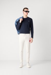 Camisa de manga larga azul marino con logo, pantalones jogger blancos con parche rojo, chaqueta de mezclilla sobre el hombro, gafas de sol negras y zapatillas blancas.