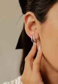 Srebrne kolczyki w kształcie okręgów z podwójną pętlą, z polerowanym wykończeniem. Zbliżenie ukazuje interakcję ucha i ręki.