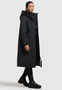 Cămașă lungă de iarnă neagră, tip puffer, cu glugă, două buzunare frontale și un design elegant. Textură netedă și închidere cu fermoar, cu accesorii decorative.