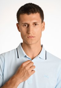 Polo de color azul claro con un cuello en contraste de color azul marino y un pequeño logo en el lado izquierdo del pecho, hecho de una tela texturizada. El modelo ajusta el cuello.