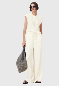 Crème-kleurige, geplooide jumpsuit met kapmouwen, een trekkoord in de taille en Wijde benen. Gecombineerd met een gestructureerde tas en zwarte sandalen.