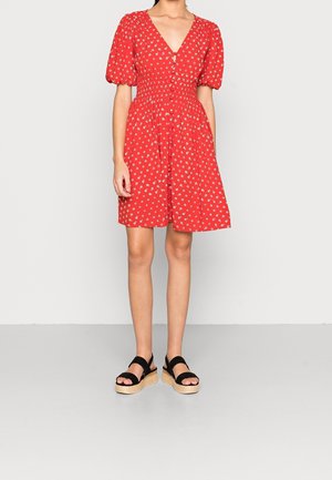 Kobieta w czerwonej sukience z krótkim rękawem, zapinanej na guziki z przodu, z białym kwiatowym wzorem i czarnych sandałach na platformie z plecioną podeszwą.