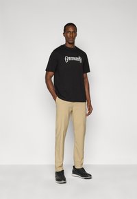 Μαύρο βαμβακερό μπλουζάκι με λευκό γραφικό σχέδιο στο μπροστινό μέρος, συνδυασμένο με μπεζ παντελόνι στενής γραμμής και μαύρα αθλητικά παπούτσια, στέκεται μπροστά σε ουδέτερο φόντο.