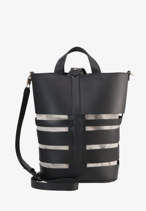 Bolso tipo cubo de cuero negro con amplias correas horizontales sobre tela beige, asa superior y correa de hombro desmontable.