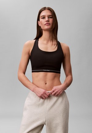 Eine Frau trägt einen schwarzen Calvin Klein Sport-BH und hellgraue Jogginghosen vor einem einfarbigen grauen Hintergrund.