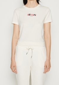 Krämfärgad t-shirt med kort ärm med en röd och svart "ARMANI EXCHANGE"-logotyp på bröstet, kombinerad med matchande ljusa sweatpants.
