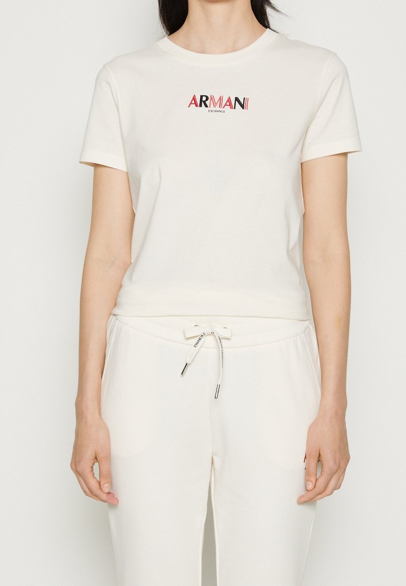 Krämfärgad t-shirt med kort ärm med en röd och svart "ARMANI EXCHANGE"-logotyp på bröstet, kombinerad med matchande ljusa sweatpants.
