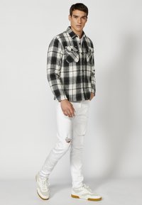 Zwart-wit geruite flanellen shirt met twee borstzakken, gecombineerd met gescheurde witte jeans en witte sneakers met gele accenten.