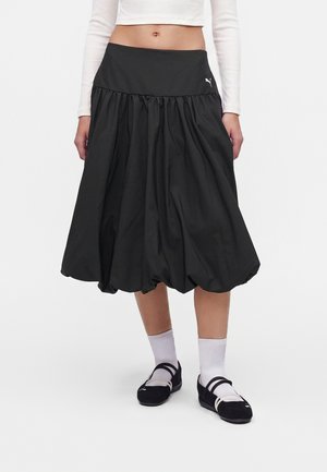 Femme portant une jupe midi noire plissée, des chaussettes blanches, des chaussures Mary Jane noires et un top court blanc à manches longues sur fond blanc.