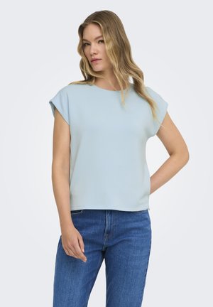 Kvinde med langt blond hår iført en lyseblå kortærmet top og blå jeans, står med den ene hånd på hoften.