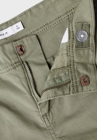 Olive green cotton pants with a zipper fly, button closure, and stitched pockets. Features a smooth texture and bronze hardware accents.