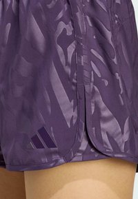 Lila Sportshorts mit einem strukturierten, abstrakten Muster, glänzender Oberfläche und einem auffälligen Adidas-Logo in der unteren linken Ecke.
