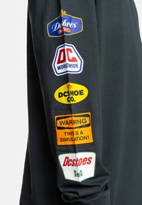 Black long-sleeve shirt featuring multiple colorful patches: blue, yellow, red, and orange logos with text, primarily on the sleeve.