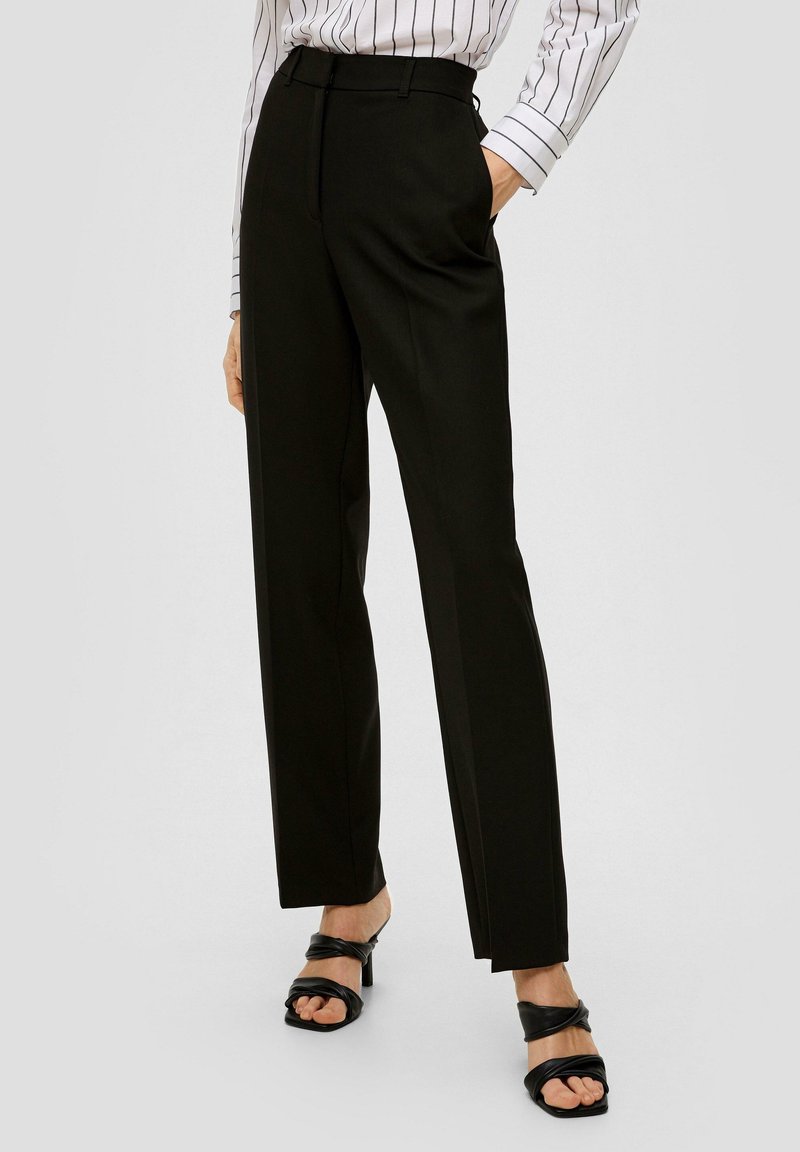Zwarte op maat gemaakte broek met een hoge taille, voorzien van scherpe plooien en zijzakken. Gemaakt in combinatie met zwarte haksandalen. Soepele stofstructuur.