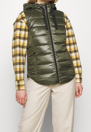 Osoba nosící olivově zelenou prošívanou vestu s kapucí přes žlutou a hnědou kostkovanou košili s dlouhým rukávem a béžové kalhoty.