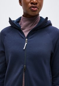 Κοντινό πλάνο ενός ατόμου που φοράει σκούρο μπλε φούτερ με φερμουάρ πάνω από ένα μοβ ζιβάγκο, το φερμουάρ είναι μερικώς ανοιχτό, ουδέτερο φόντο.