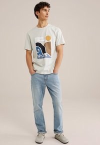 Světle šedé bavlněné tričko s barevným abstraktním motivem vln a slunce. Spárováno se světle modrými džíny a šedými nazouvacími botami.