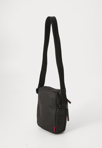 Black crossbody bag with a textured fabric, square shape, adjustable black strap, and red accent tag. Features a top zipper closure.