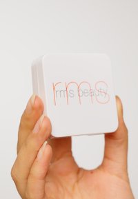 Contenitore quadrato bianco etichettato "rms beauty" in testo grigio e arancione. Tenuto in mano, con superfici lisce e bordi arrotondati.