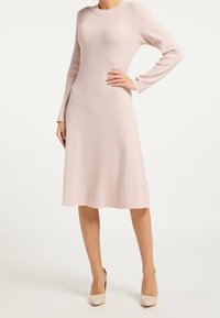 Knädjup, långärmad klänning i mjukt rosa stickat tyg. Har rund halsringning och en lätt utsvängning i fållen, kompletterad med beige klackskor.