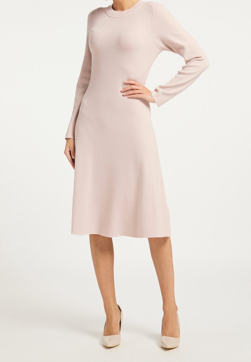 Knädjup, långärmad klänning i mjukt rosa stickat tyg. Har rund halsringning och en lätt utsvängning i fållen, kompletterad med beige klackskor.