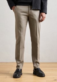 Beiget räätälöidyt housut, joissa on sileä pinta, kapea istuvuus ja hillityt sivusaumat, yhdistettynä mustiin slip-on-kenkiin, joissa on paksu pohja.