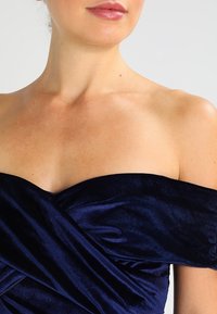 Mörkblå sammet off-shoulder topp med en draperad design och slät textur samt mjukt glans. Fokusera på halsringningen och axelns detaljer.