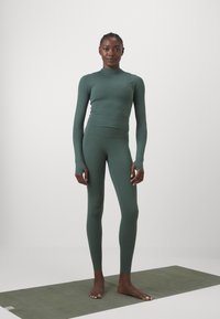 Das grüne Activewear-Set besteht aus einem langärmligen Oberteil und hochgeschnittenen Leggings, hergestellt aus dehnbarem Material, mit nahtlosem Design und taillierter Silhouette.