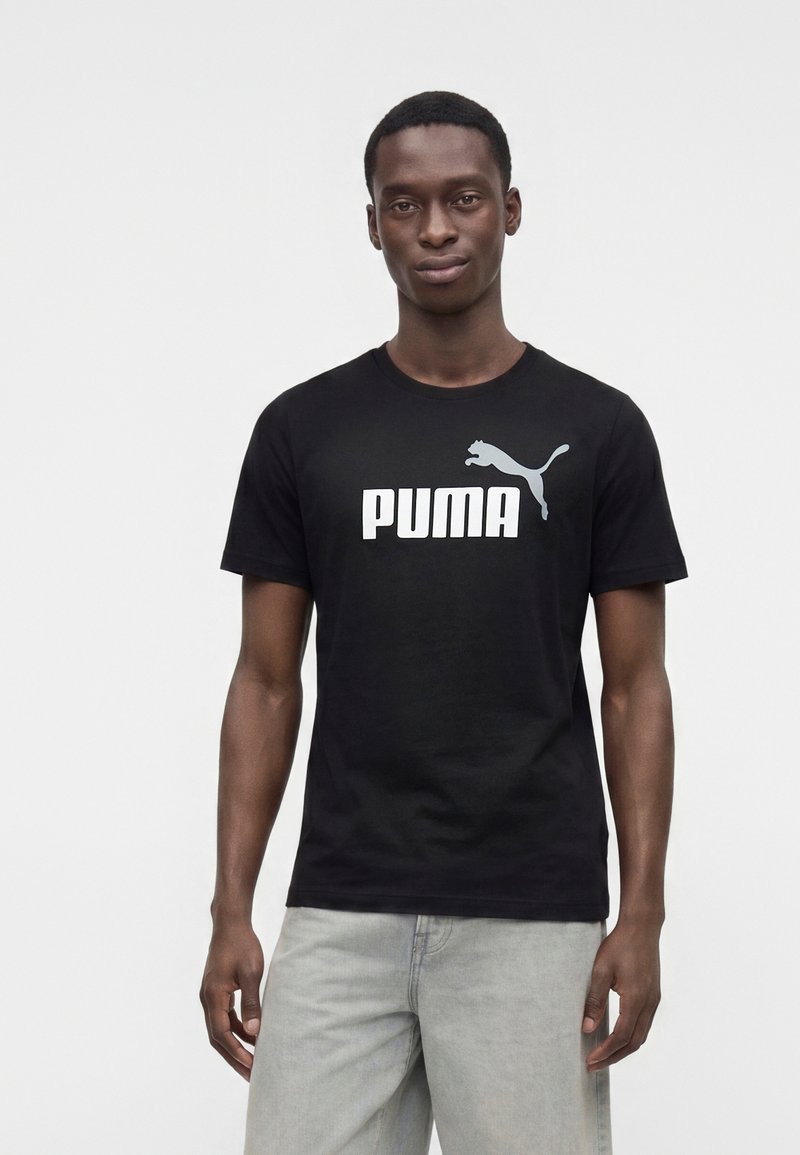 Jeune homme portant un t-shirt noir Puma avec logo et un jean gris clair, debout devant un fond clair uni.