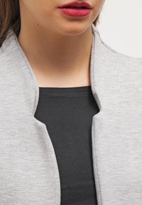 Szara, teksturowana kurtka z kołnierzem, nałożona na czarną bluzkę. Skupienie na dekolcie i czystych liniach projektu.