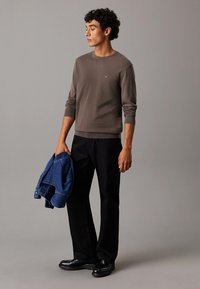 Maglione di maglia marrone, pantaloni neri e scarpe nere. Tiene in mano un giubbotto di denim blu. Design semplice con polsini e fondo a coste.