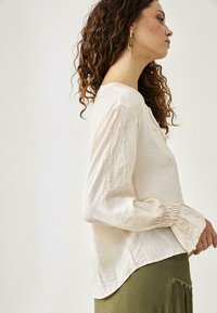 Blusa color crema con mangas largas, que presenta delicados bordados en las mangas y puños fruncidos. Tela suave y ligera.