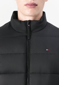 Jachetă puffer neagră cu guler înalt și închidere cu fermoar, având cusături orizontale și un mic logo în roșu, alb și albastru.