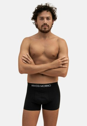 Homme torse nu avec des cheveux bouclés et une barbe, se tient les bras croisés, portant des boxers noirs avec une ceinture "MASSI MORINO".