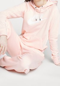 Ljusrosa hoodie och sweatpants-set gjort av mjukt material, med en vit Nike-logotyp, dragskon och avslappnad passform med samlade mudd.