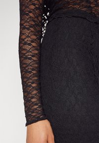 Top de encaje negro con mangas largas, que presenta un patrón floral. Tejido texturizado con bordes con forma de onda en el escote y el dobladillo de las mangas.