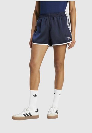 Dunkelblaue Shorts mit weißem Besatz, ausgestattet mit einem elastischen Bund und drei Streifen an den Seiten. Kombiniert mit weißen Schuhen und Socken mit Logo.