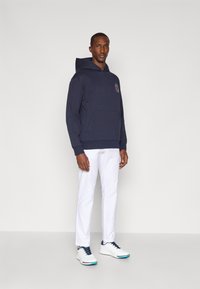 Marine blå hettegenser med frontlomme, ribbede mansjetter og logo detalj på brystet, kombinert med hvite bukser og hvite joggesko.