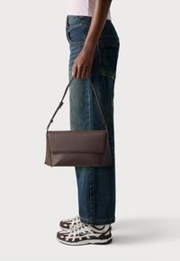 Sac à main en cuir marron avec une forme structurée et une poignée supérieure, tenu par une personne portant un jean bleu et des baskets marron.