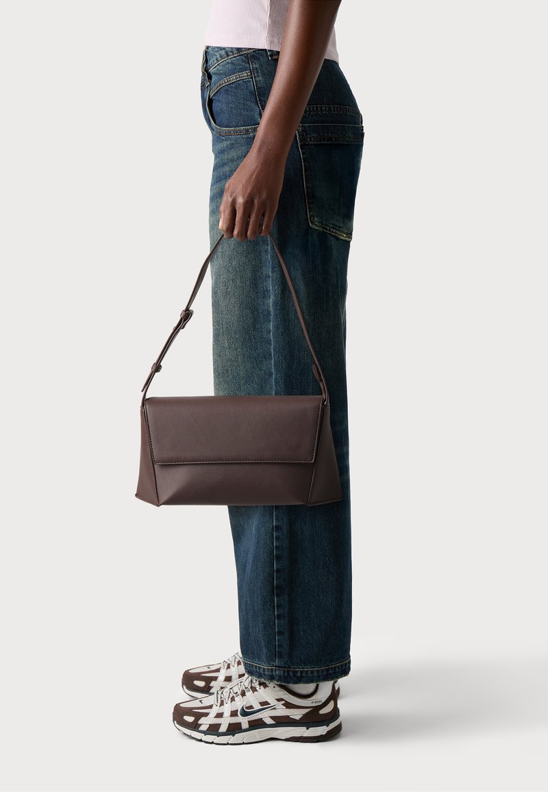 Sac à main en cuir marron avec une forme structurée et une poignée supérieure, tenu par une personne portant un jean bleu et des baskets marron.