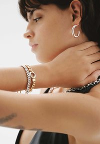 Pulseras de cadena de oro y plata en un brazo, junto a un gran pendiente de aro de plata curvado. La piel tiene una textura suave y un tono neutro.