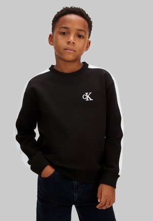 Junger Junge mit kurzen lockigen Haaren, trägt schwarzen Sweatshirt mit weißen Streifen an den Ärmeln und "cK"-Logo auf der Brust, Hände in den Taschen.