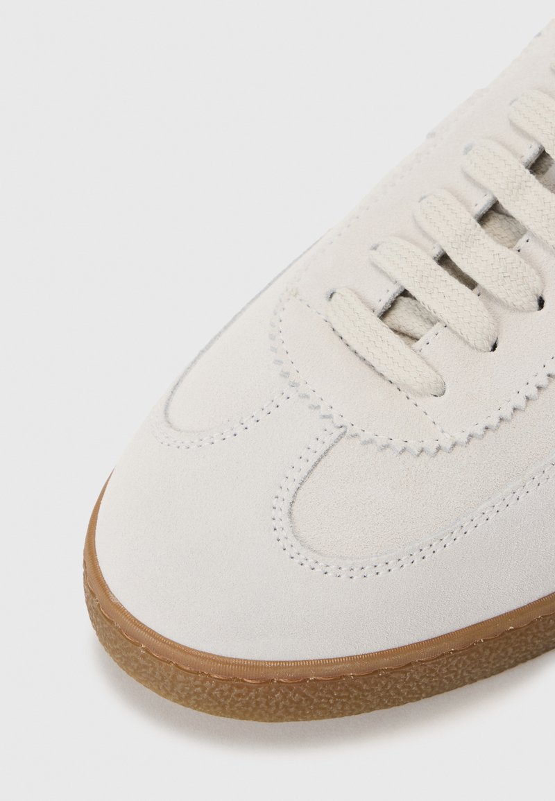 Sneaker en daim gris clair avec un bout arrondi, une semelle en caoutchouc texturée et cinq lacets symétriques. Les accents de couture mettent en valeur les détails du design.