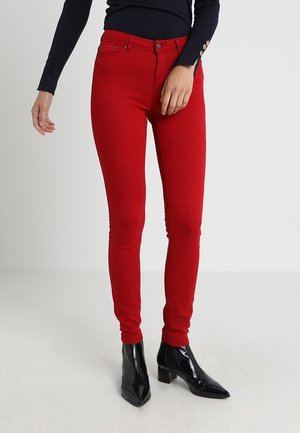 Czerwone obcisłe spodnie noszone z czarną bluzką z długim rękawem i czarnymi botkami z ostrym noskiem, stojące na jednolitym jasnym tle.