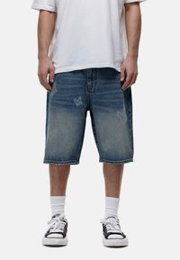 Shorts in denim di un blu chiaro con una finitura sbiadita. Vestibilità rilassata, lunghezza al ginocchio, con una cucitura centrale e delicati motivi a farfalla.
