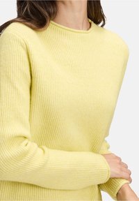 Femme portant un pull en maille côtelée jaune douce à manches longues, ajustant une manche près du poignet, sur un fond uni.